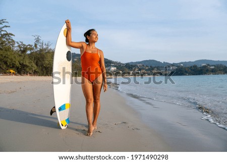 Portrait of Attractive Asian woman in swimwear holding surfboard and walking on tropical beach at summer sunset. Confidence female relax and enjoy outdoor activity lifestyle and extreme sports surfing