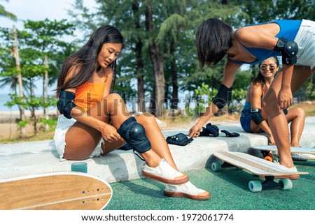 Asian woman with girl friends wearing safety skateboarding knee pad for skating at skateboard park. Happy female friendship enjoy summer outdoor active lifestyle play extreme sport surf skate together
