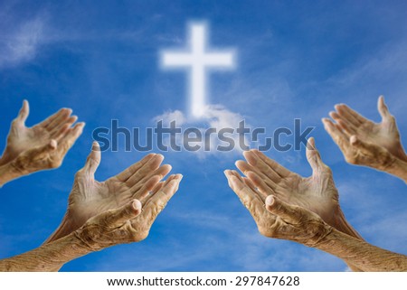 The old woman\'s hands praying over blurred the cross on the sky background.