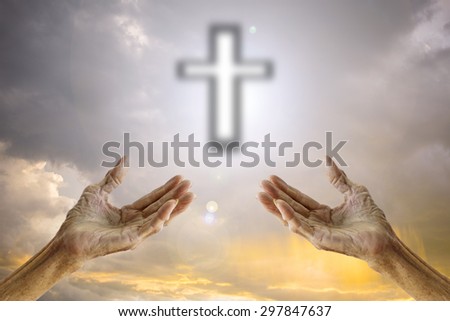 The old woman\'s hands praying over blurred the cross on the sky background.