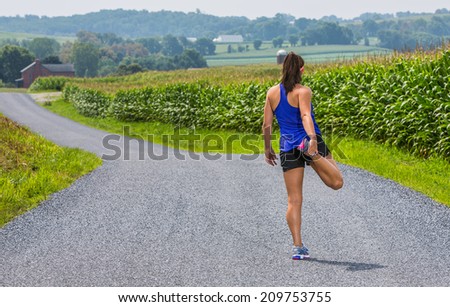 Young fit female athlete getting ready to run by stretching on a country road.