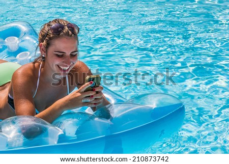 Young female adult lying on raft and checking her phone having fun in swimming pool during summer.