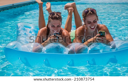 Two young female adults lying on raft in swimming pool checking their cell phones and having fun during summer.