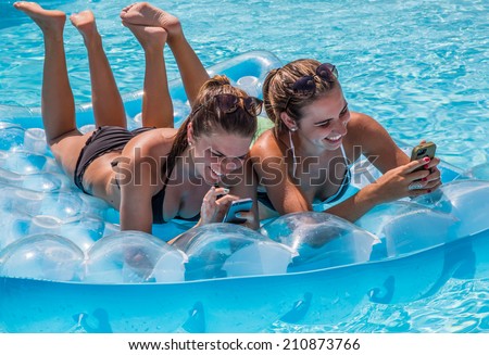 Young female adults lying on raft checking their phones and having fun in swimming pool during summer.