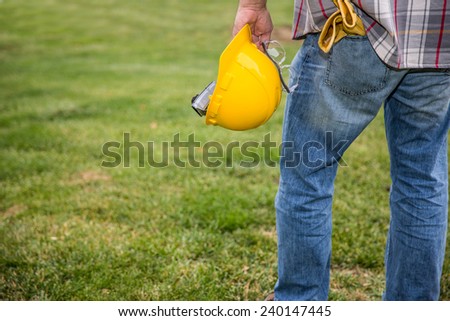Man with work gloves in pocket and a construction helmet and safety glasses in hand against a grass background.