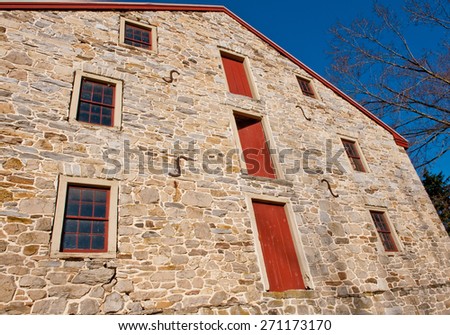 Wide angle view of side of stone barn building with red doors and windows against blue sky.