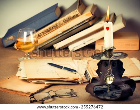 Vintage pocket watch, book, pen, candlestick with burning candle made from lether and old glasses on a table with books and glass of wiskey on background