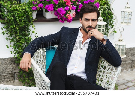 A beautiful man in a jacket and shirt sitting on a chair and looking at the camera, green and flowers, outside