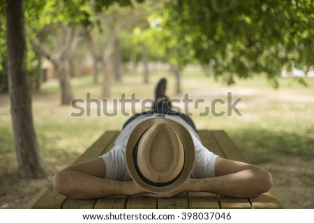 Restful young man wearing a straw hat laying down on a wooden table in the middle of the forest at a park
