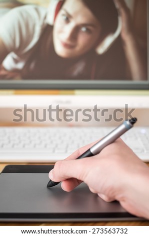 First person view for graphic tablet and hand with pen. In the background is the blurred keyboard and LCD monitor with retouched photo. Selective focus on pen.