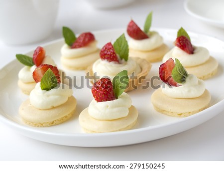 Collection of small cakes with whipped cream, fruits, mint on white plate on table against background
