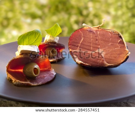 Smoked dried gammon ham and starters (sandwich) with olives, cheese and basil on brown platter against green background
