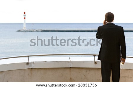 A business man standing on a balcony and talking on the mobile phone with a glass of champagne in his hand while looking at the sea with a lighthouse on the horizon