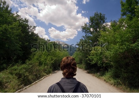 A young man walking forward towards a lone path surrounded by woods under a sunny and cloudy sky