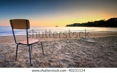 A broken and old chair left on the sand at the beach pointed to the sunset behind a hill. The concept of loneliness and missing