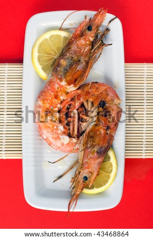 A rectangular white dish on wooden sticks and red background with grilled argentinian prawns and lemon.