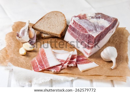 Salty bacon with garlic, black rye bread and black pepper.. Slices of bacon on the parchment on a white wooden table