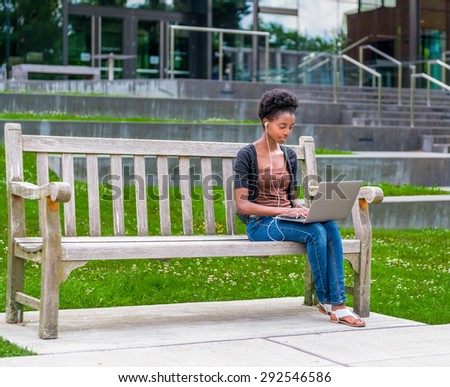 Young African American student sitting on bench doing homework while listening to music
