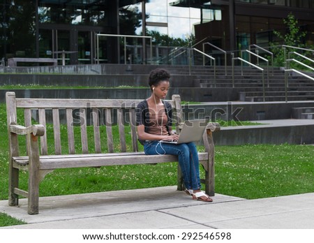 African American college student sitting on bench doing homework while listening to music.