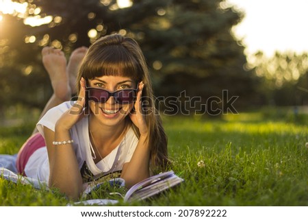 Beautiful smiling  young woman lying on grass and reading blue book, summer green park. Beautiful smiling  young woman with sunglassses lying on grass and reading blue book, summer green park.