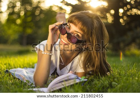 Beautiful smiling  young woman with sunglassses lying on grass and reading blue book, summer green park. Female student girl outside in park. Happy young university student