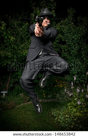 A man in an old style suit, hat and sunglasses jumps while pointing a gun.