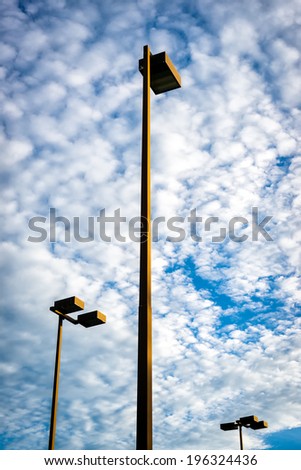 Three very tall lights, or lamps, of the kind to illuminate parking lots or park grounds stand against a cloudy sky.