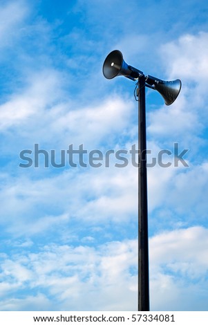 A public address system speakers tower against a cloudy, blue, daytime sky.