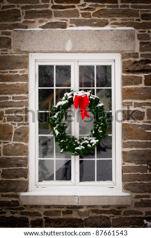 A green spruce Christmas wreath with red ribbon hangs in the middle of an old, snow covered window pane set in stone wall.