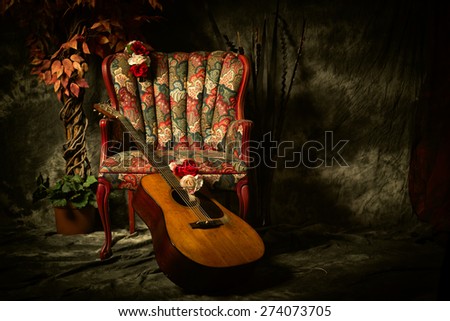 A vintage acoustic guitar leans against an empty, antique patterned armchair. Shot in chiaroscuro style lighting with room for your copy.