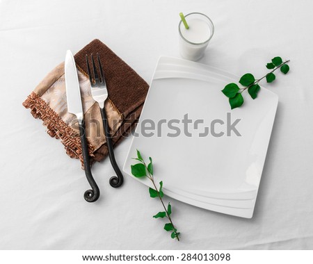 A clean, white fancy plate lies empty on a white background cloth in a simple, beautiful place setting with knife and fork, sprigs of leaves and small drink.