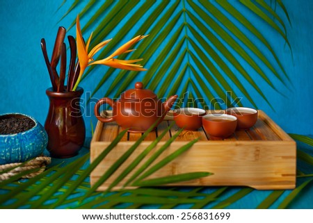 Handmade coconut bowl with tea, set of wooden spoons, yellow flower teapot and three teacups on a wooden box with green leaves on blue wooden background.