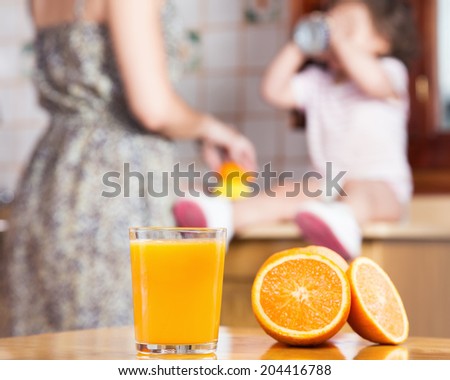 A freshly squeezed orange juice in a glass with oranges on a wood table. In the background a women is squeezing juice with a juicer and a baby girl is drinking the juice.