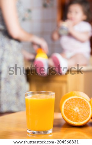 A freshly squeezed orange juice in a glass with oranges on a wood table. In the background a women is pressing juice with a juicer and a baby girl is drinking the juice.