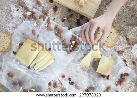 Woman hand holding a piece of cheese of snack of cheese, roasted almonds and toast. Rustic lunch of brown on stone table in the countryside