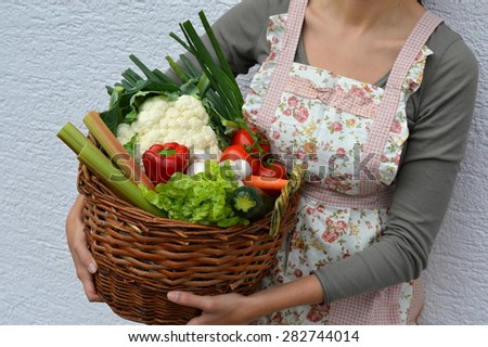 A woman wearing a cooking apron is showing a wicker basket full of fresh vegetables