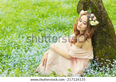 beautiful sexy girl with red hair with flowers in her hair sitting near a tree in a pink dress in the meadow with blue flowers