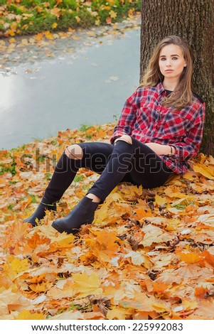 beautiful charming young attractive girl with large blue eyes, with long dark hair in the autumn forest sits on the leaves near the water in black pants and shirt