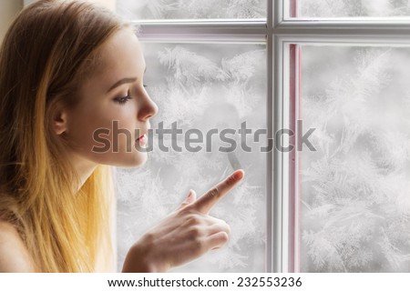 beautiful young girl sitting by the window winter day and draws the sun on the frozen window