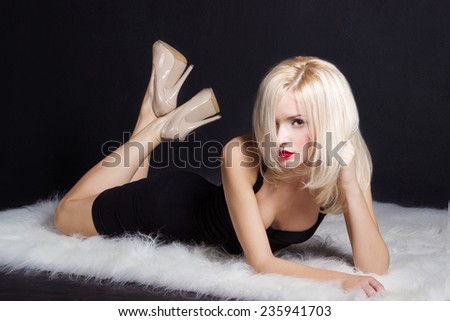 beautiful sexy elegant striking blonde woman with bright makeup red lips in a black dress lies on the white fur in the Studio on a black background