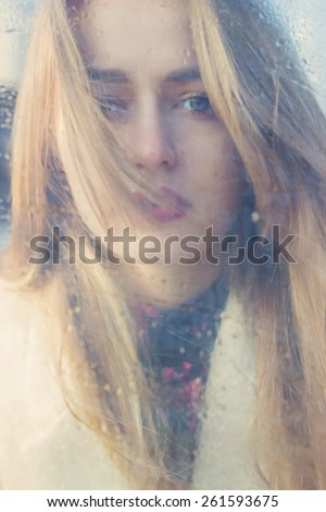 beautiful cute tender young girl with big sad eyes with white hair and dark lipstick in a bright coat looks into the camera through wet glass