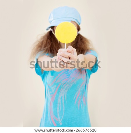 girl in blue shirt and cap with two tails holds in front of a big yellow candy