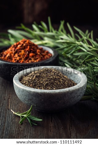 Ground black pepper in stone bowl, chili flakes and rosemary in background