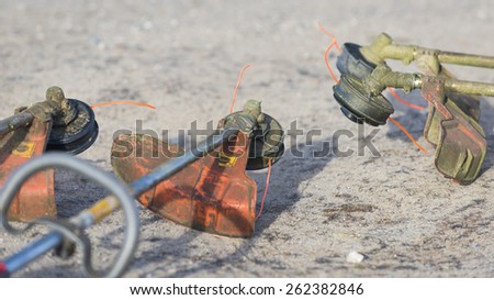 Four well used weed trimmers laying in a semicircle on a sandy road. Dirty orange trimmer guards and heads covered with grass clippings. Trimmer string is fluorescent orange.