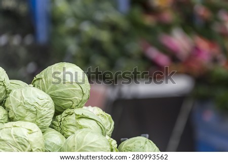 Heads of green cabbage pile up in a farmer\'s market stall.  Other vegetables stacked on tables out of focus in the background.