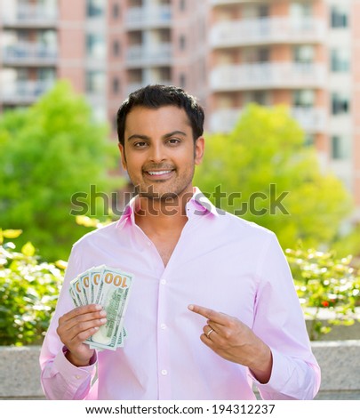 Closeup portrait, super happy excited successful young business man pointing to money dollar bills in hand, isolated background of trees, building. Financial reward