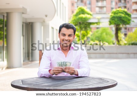 Closeup portrait, super happy excited successful young business man holding money dollar bills in hand, isolated background of trees, building. Financial reward