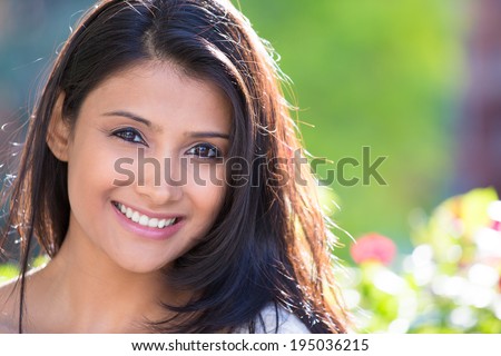 Closeup headshot portrait of confident smiling happy pretty young woman, isolated background of blurred trees, flowers. Positive human emotion facial expression feelings, attitude, perception