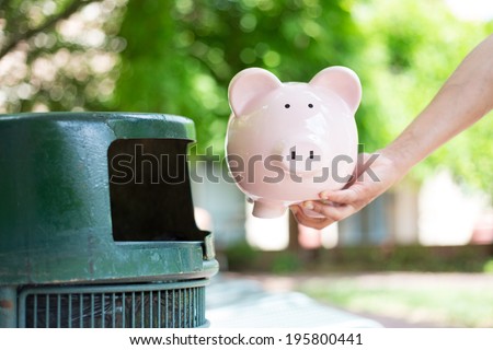 Closeup cropped portrait of someone hand tossing piggy bank into trash can, isolated outdoors green trees background. Bad financial investment decisions concept