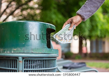 Closeup cropped portrait of someone hand tossing cash dollar bills money, hundred dollar bills in trash can, isolated outdoors green trees background. Bad financial investment decisions concept